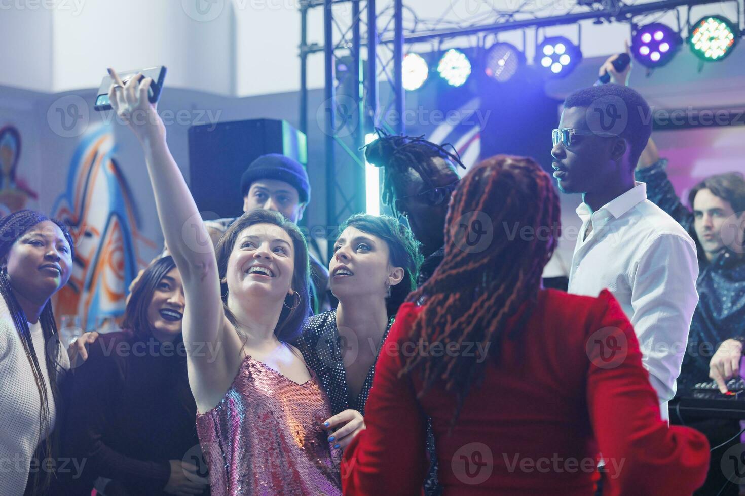 vrolijk clubbezoekers nemen groep foto Aan smartphone terwijl feesten Aan druk nachtclub dansvloer met lichten. zorgeloos glimlachen vrouw maken mobiel telefoon selfie met vrienden in club