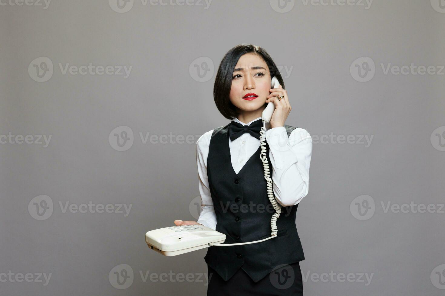 jong zelfverzekerd Aziatisch serveerster in uniform pratend Aan vaste telefoon telefoon en op zoek weg. echt vrouw receptioniste Holding retro telefoon en sprekend met restaurant personeel foto