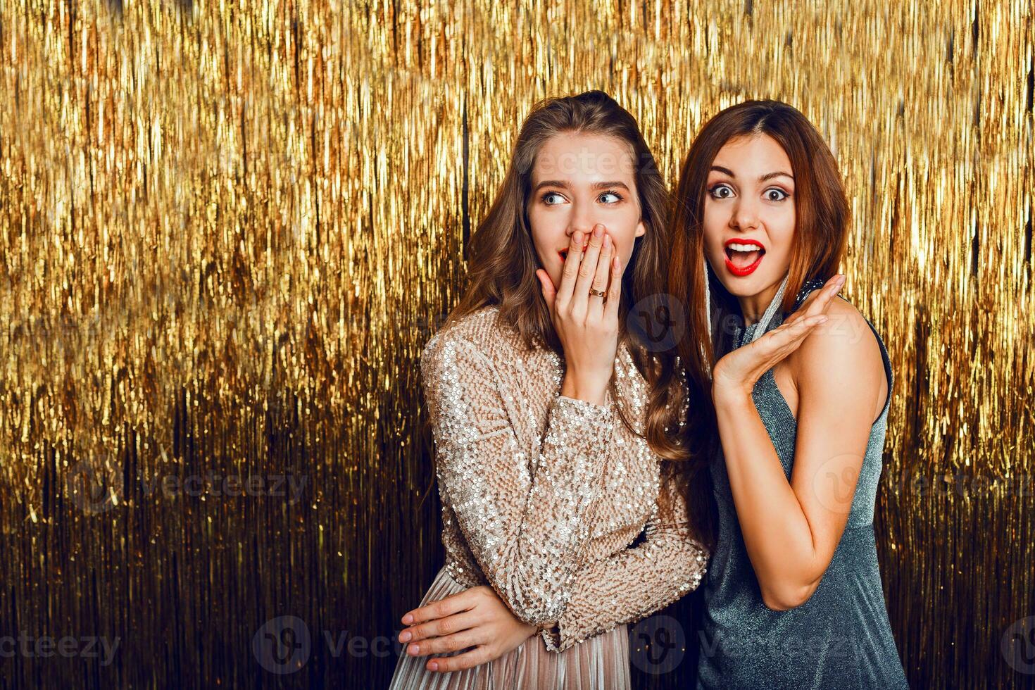 dichtbij omhoog studio beeld van twee verbazingwekkend sexy vieren meisjes met rood lippen, verrassing gezicht, lachend , poseren Aan gouden sparkly achtergrond. nieuw jaar partij humeur. foto