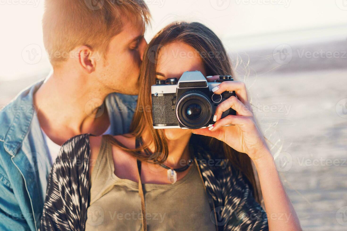 paar in liefde poseren Aan de avond strand, jong hipster meisje en haar knap vriendje nemen foto's met retro film camera. zonsondergang warm licht. foto