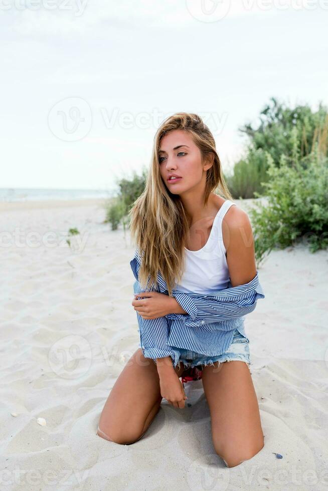 dichtbij omhoog portret van blond verleidelijk meisje met lang glimmend golvend haren Aan zomer strand genieten van vakantie. gestreept blouse, jeans korte broek. rietje hoed. foto