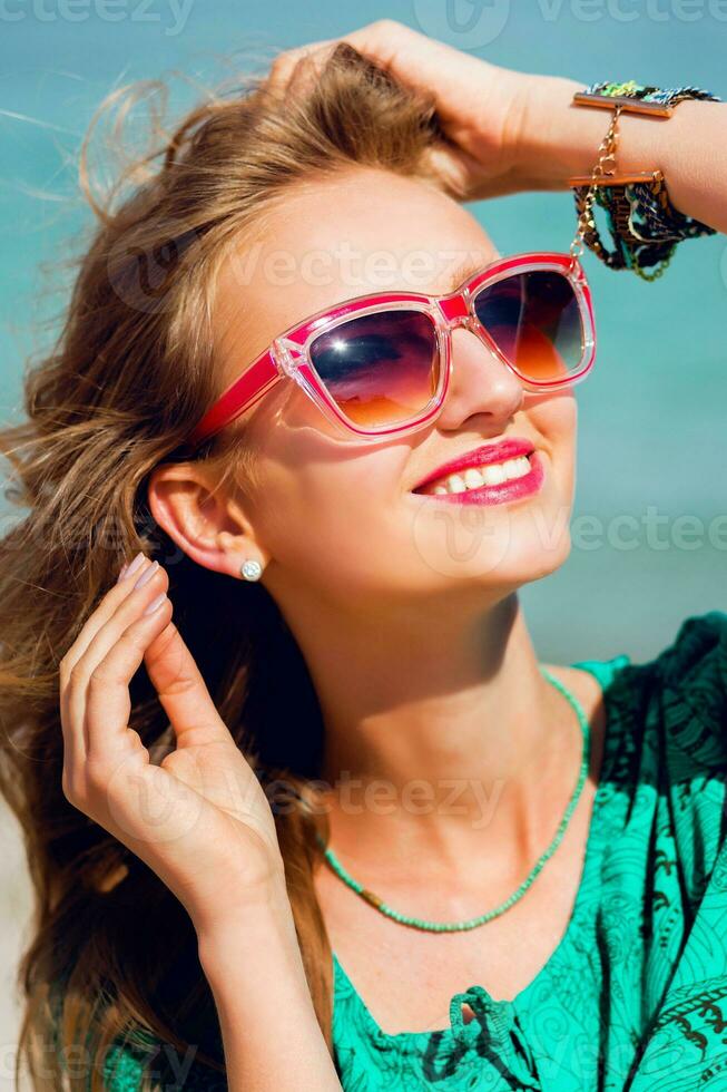 buitenshuis zomer portret van mooi jong blond mooi vrouw in koel zonnebril poseren Aan de zonnig tropisch strand. vervelend elegant strand kleding. foto