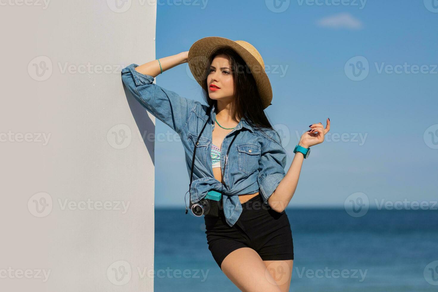 verbijsterend vrouw met perfect figuur in sexy shorts en rietje hoed poseren Aan de strand. blauw lucht en zee Aan achtergrond. foto