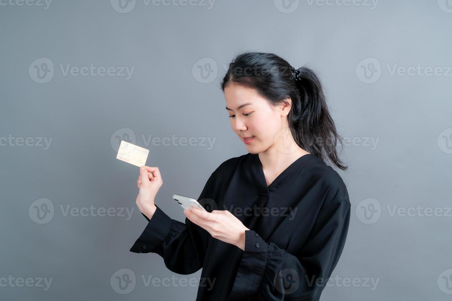 portret van een gelukkig jong Aziatisch meisje dat een plastic creditcard toont terwijl ze een mobiele telefoon op een grijze achtergrond houdt foto