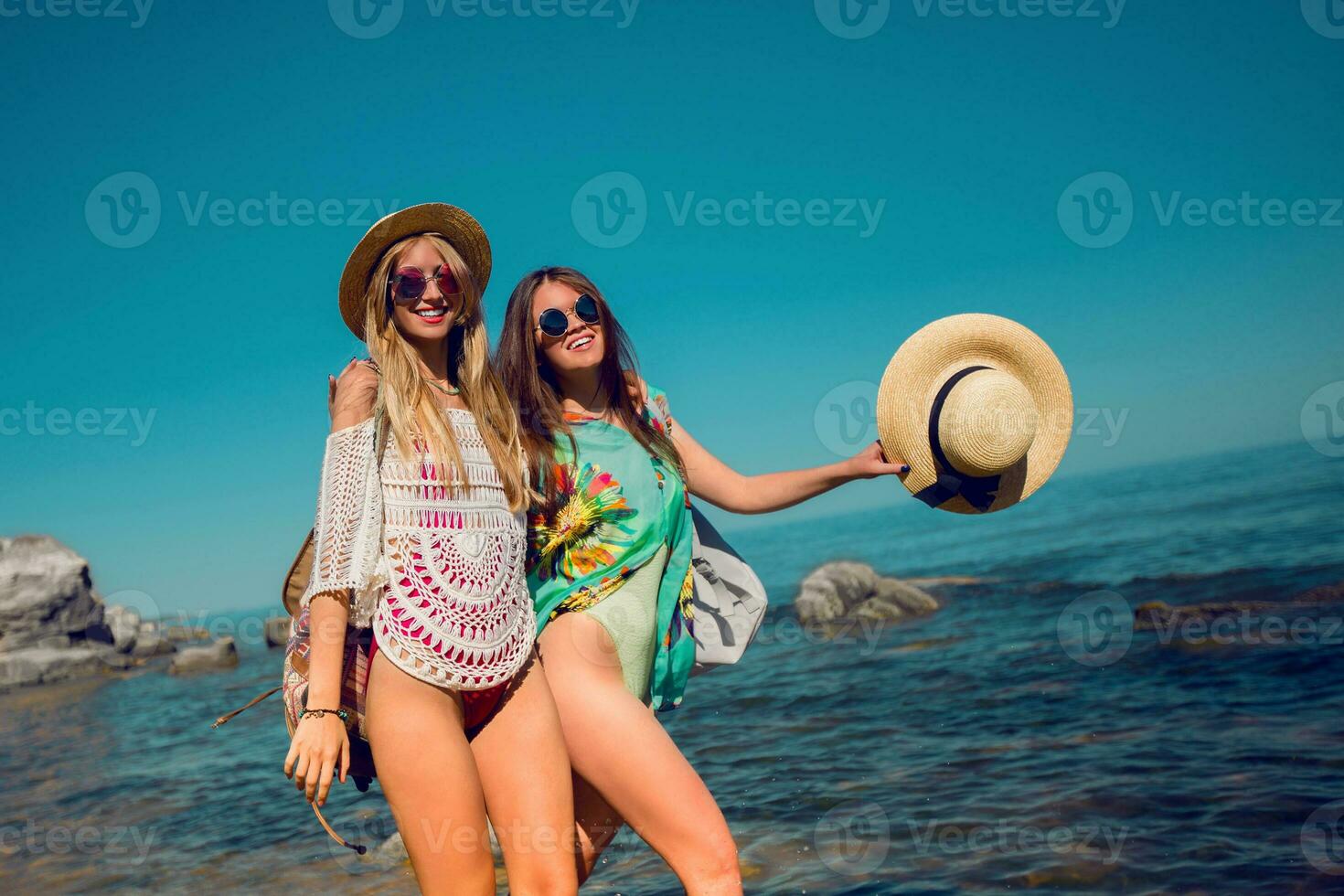 twee mooi vrienden wandelen in de strand en hebben plezier. meisjes vervelend elegant zwemkleding , rietje hoed en koel zonnebril. zomer helder kleuren. foto