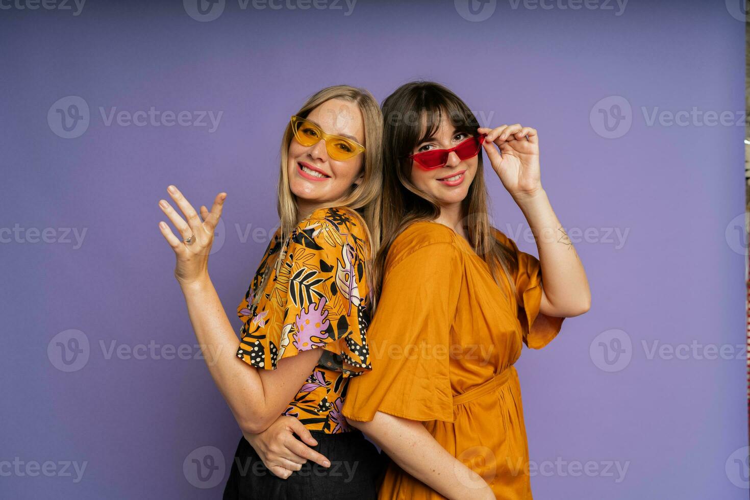 dichtbij omhoog portret van twee elegant Dames in zonnebril en modieus zomer kleren poseren Aan Purper bakgrond in studio. foto