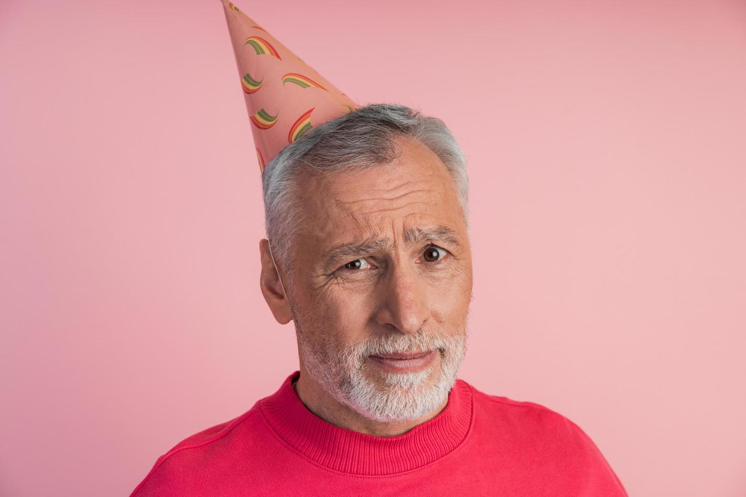 verdrietig, boos senior man in een feestelijke hoed op een roze achtergrond. foto