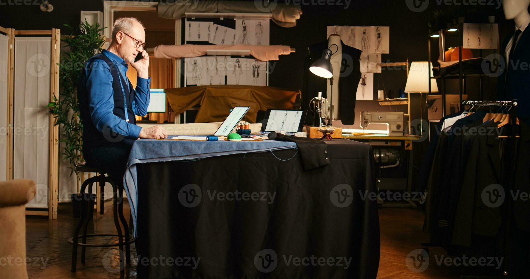 vakman roeping cliënt in werkplaats, zorgen voor perfect metingen en kleding stof Aan mobiel telefoon telefoongesprek. professioneel couturier ontwerpen handgemaakt kleren en ambacht op maat gemaakt artikelen. handheld schot. foto