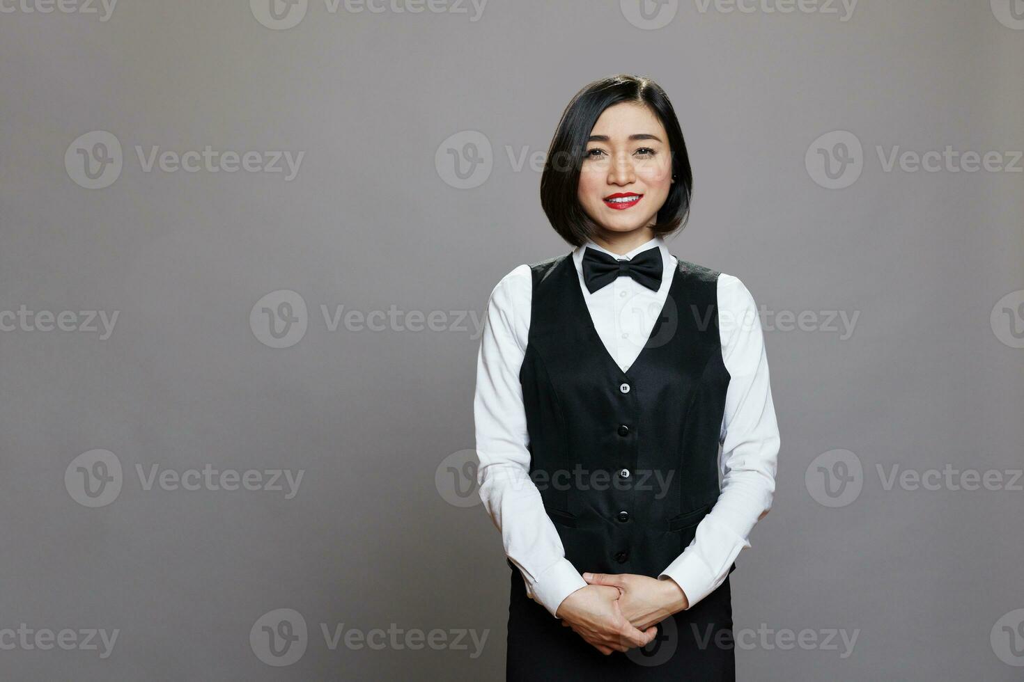 glimlachen aantrekkelijk Aziatisch serveerster vervelend uniform met boog stropdas staand en op zoek Bij camera. restaurant professioneel receptioniste poseren voor studio portret Aan grijs achtergrond foto