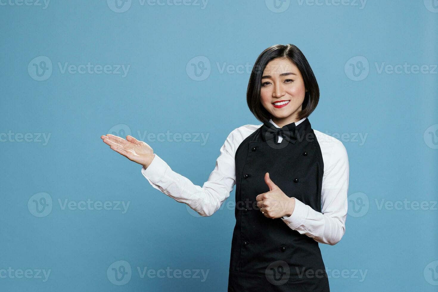 vrolijk Aziatisch vrouw receptioniste tonen duim omhoog en richten naar kant met hand- portret. serveerster weergeven tevredenheid gebaar en presentatie van naar Rechtsaf richting terwijl op zoek Bij camera foto