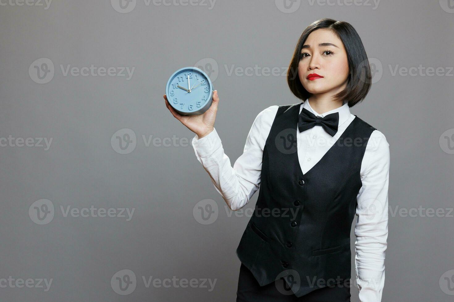 hotel vrouw receptioniste Holding retro alarm klok en op zoek Bij camera met echt gelaats uitdrukking. jong Aziatisch serveerster vervelend uniform tonen tijd beheer concept portret foto