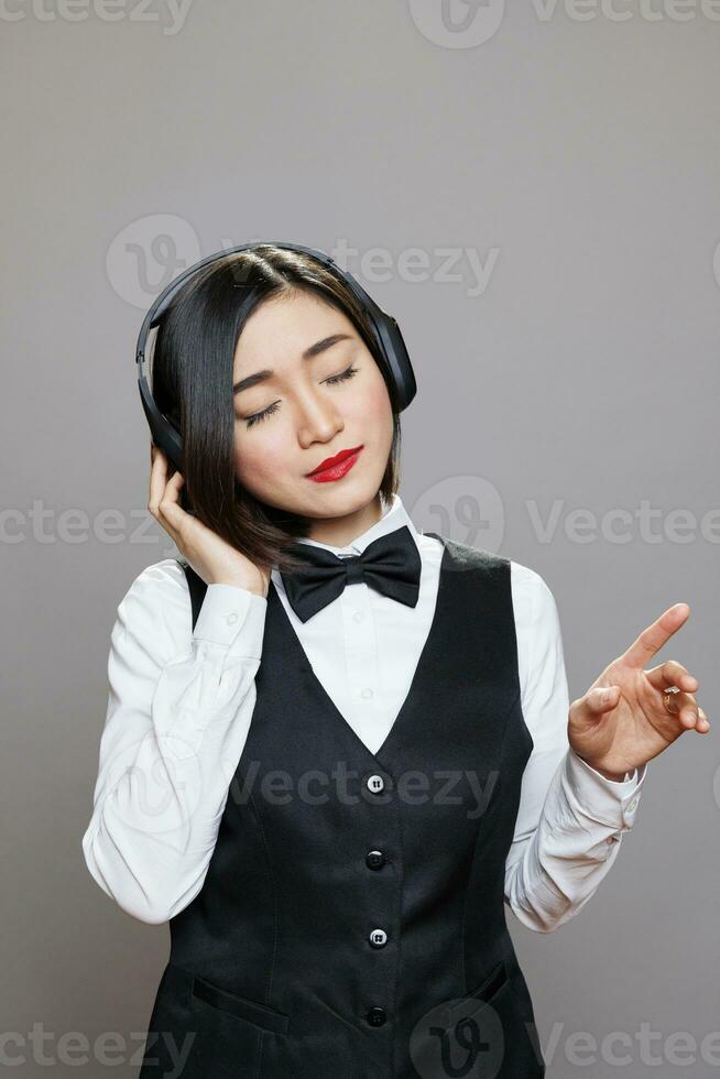 Aziatisch vrouw receptioniste in uniform genieten van afspeellijst in koptelefoon. jong aantrekkelijk serveerster met Gesloten ogen luisteren naar muziek- in draadloze oortelefoons terwijl poseren in studio foto