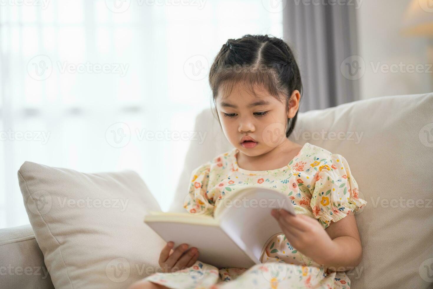 blij gelukkig Aziatisch kind baby meisje glimlachen en lezing boek terwijl zittend Aan bankstel sofa in leven kamer Bij huis. meisje relex lezing boek glimlach Bij sofa in de huis. terug naar school- concept. foto