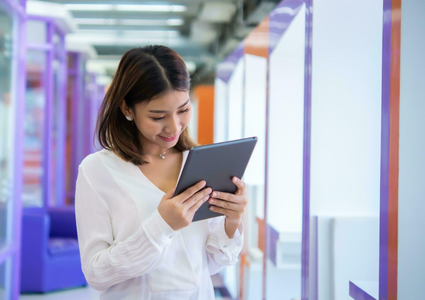 mooi Aziatisch bedrijf vrouw is staand en op zoek naar beneden Bij haar tablet met een gelukkig uitdrukking, digitaal marketing. foto