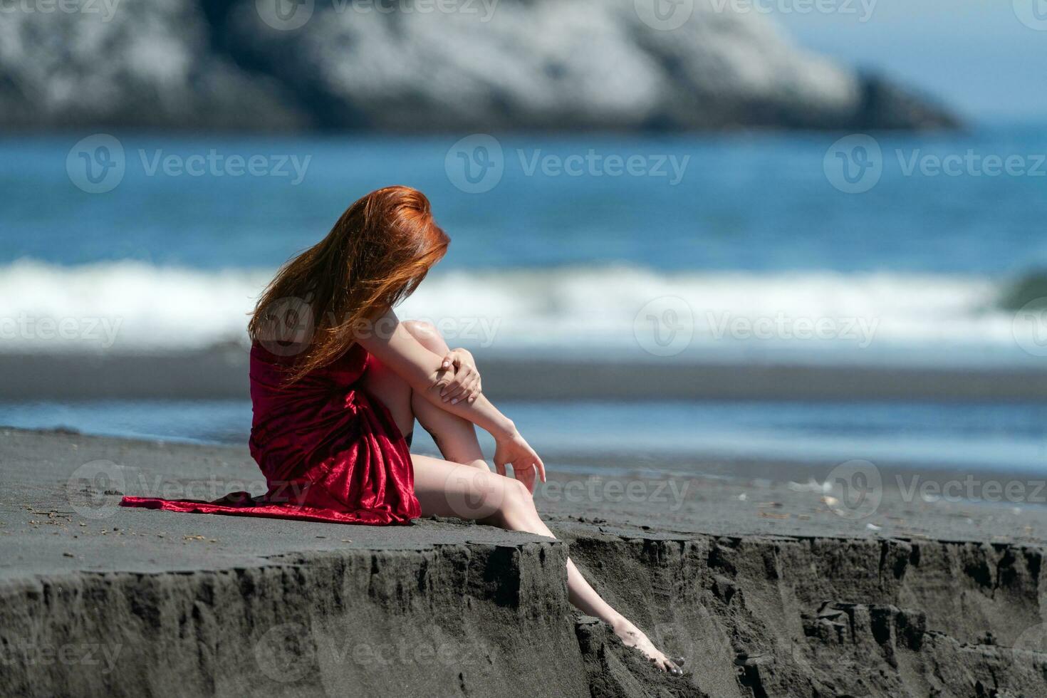 onherkenbaar vrouw in rood jurk met verheven zoom zittend Aan strand ontspanning gedurende strand vakanties foto