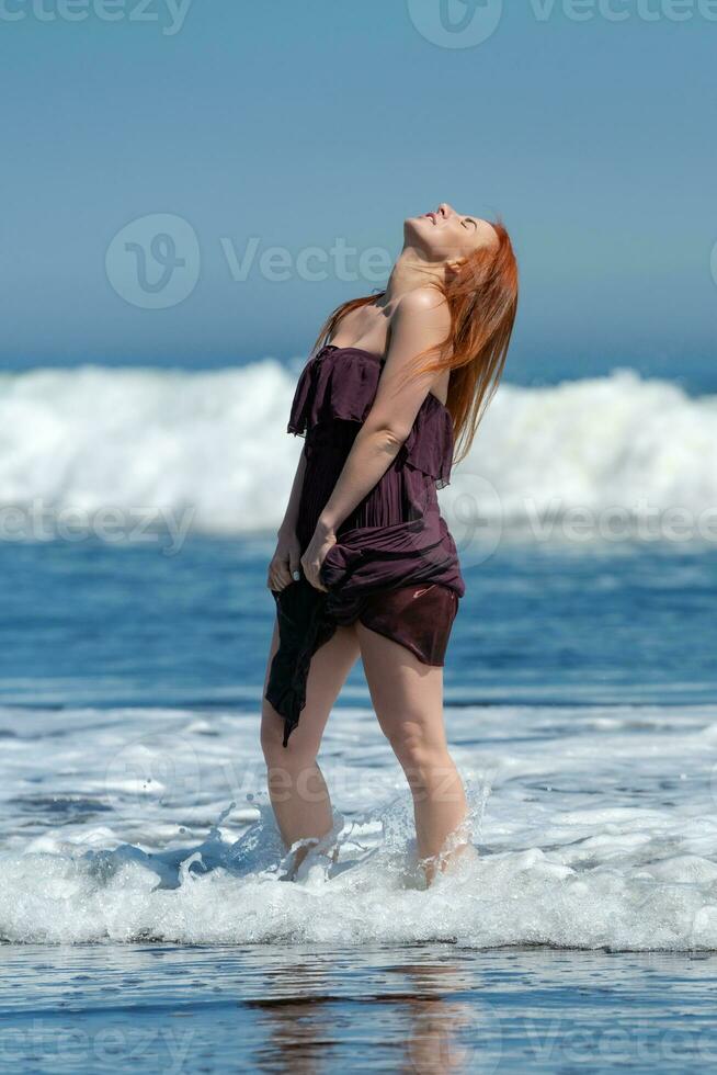 gelukkig vrouw in lang jurk met verheven zoom staand enkel diep in water Aan strand, hoofd op zoek naar boven foto