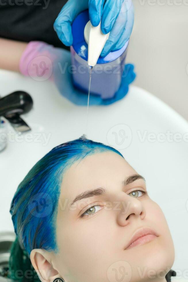 detailopname visie van kapper knijpt shampoo van buis in hoofd van vrouw met blauw haar- terwijl het wassen haar- in speciaal wastafel foto