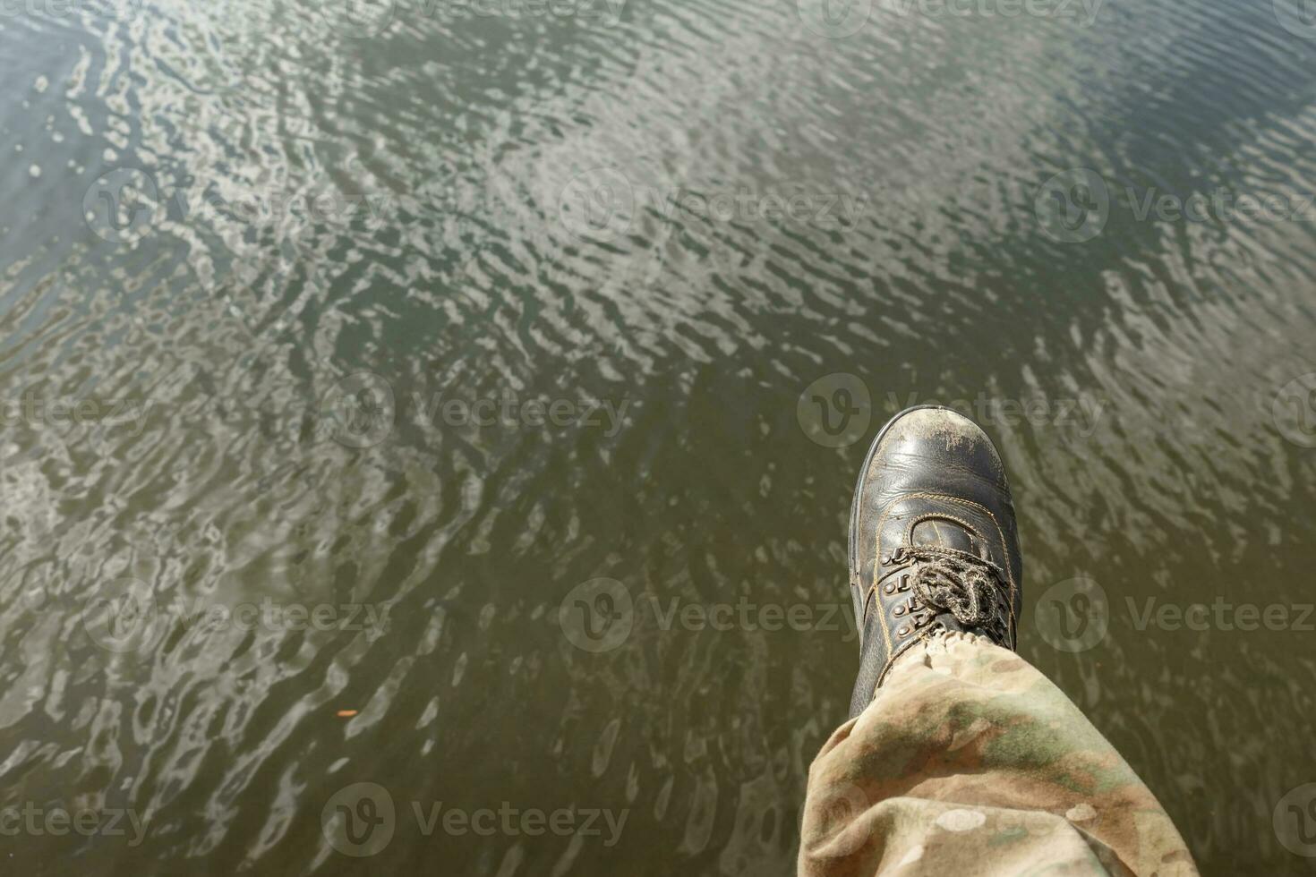 Mannen poten in camouflage broek en oud laarzen bengelen bovenstaand de water. foto