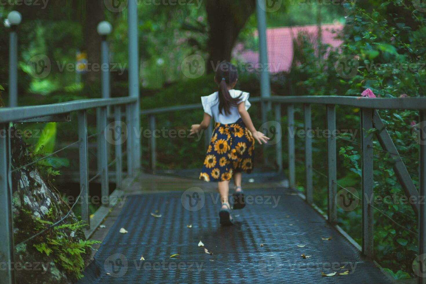 schattig weinig meisje wandelen Aan een staal brug in een botanisch tuin met groen planten en kleurrijk bloemen in de omgeving van. kinderen aan het studeren natuur foto