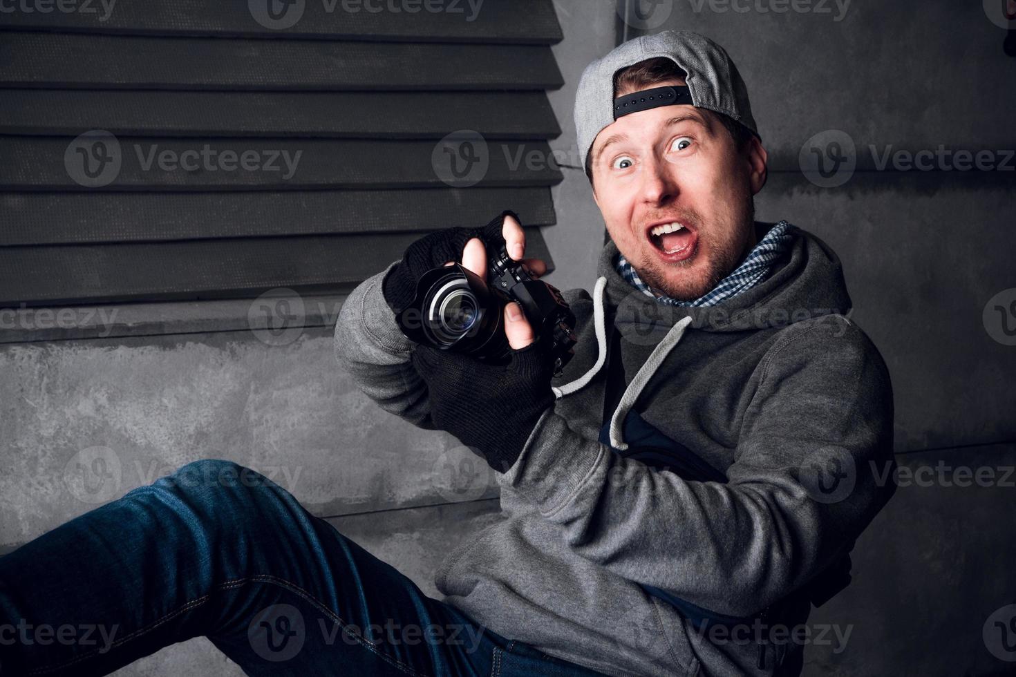 man met een camera en een grappig gezicht zittend op een stoel in een grijze studio foto