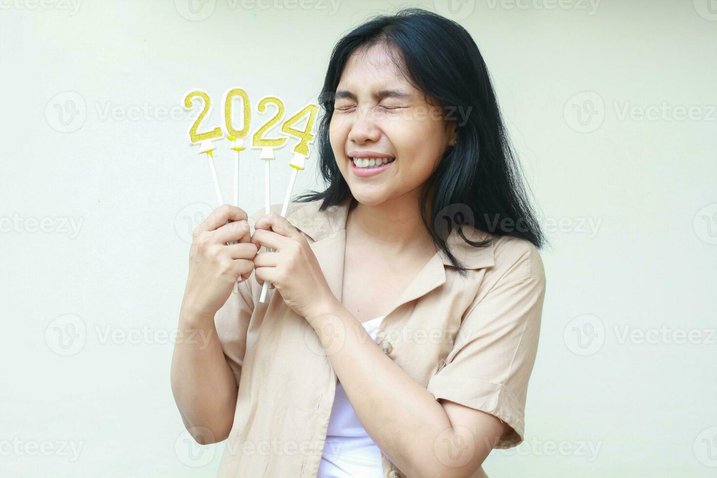 opgewonden Aziatisch jong vrouw vieren nieuw jaren vooravond door Holding gouden kaarsen getallen 2024 met Gesloten oog vervelend gewoontjes bruin overhemd geïsoleerd Aan wit achtergrond foto