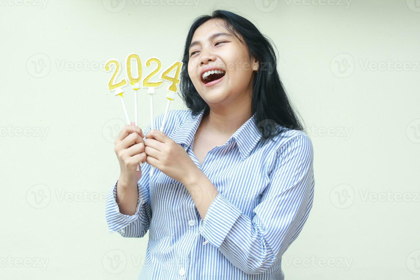 portret van gelukkig Aziatisch jong vrouw collega glimlachen en op zoek terzijde naar 2024 figuur kaarsen houden Aan handen vervelend blauw strepen gewoontjes overhemd naar vieren nieuw jaren vooravond, geïsoleerd Aan wit foto
