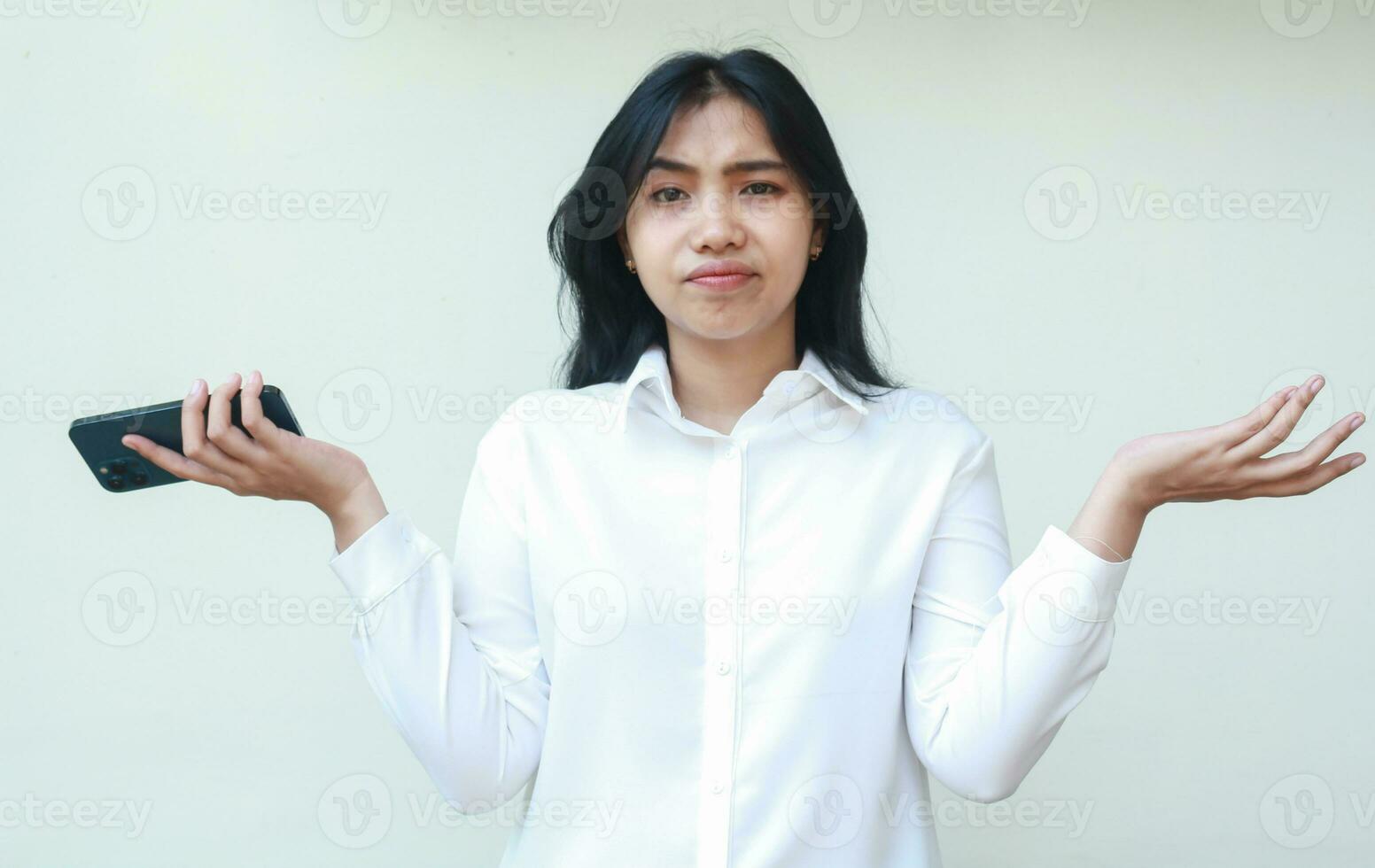 portret van verward Aziatisch vrouw secretaris Holding smartphone schouderophalend schouders houden palm zijwaarts op zoek Bij camera met onzeker uitdrukking, hebben Nee idee gebaar, vervelend wit formeel pak, staand foto