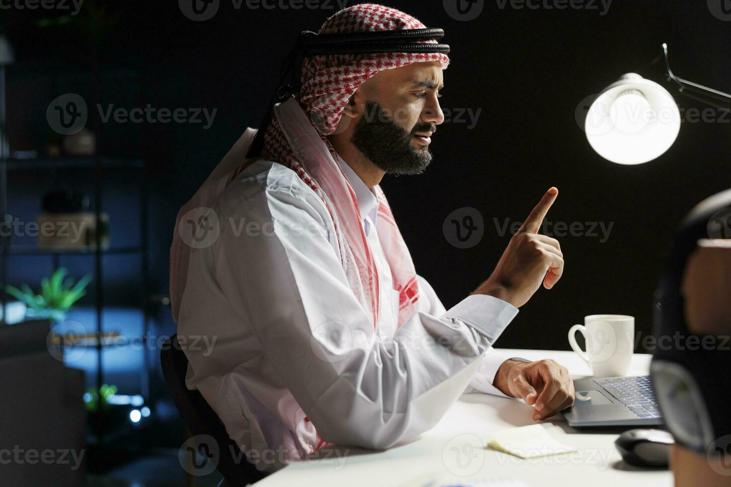 zijaanzicht schot van Islamitisch vent vervelend traditioneel kleding gezeten Bij zijn bureau hebben een conferentie telefoontje Aan zijn laptop. beeld shows Arabisch freelancer verdiept in een gesprek Aan zijn persoonlijk computer. foto