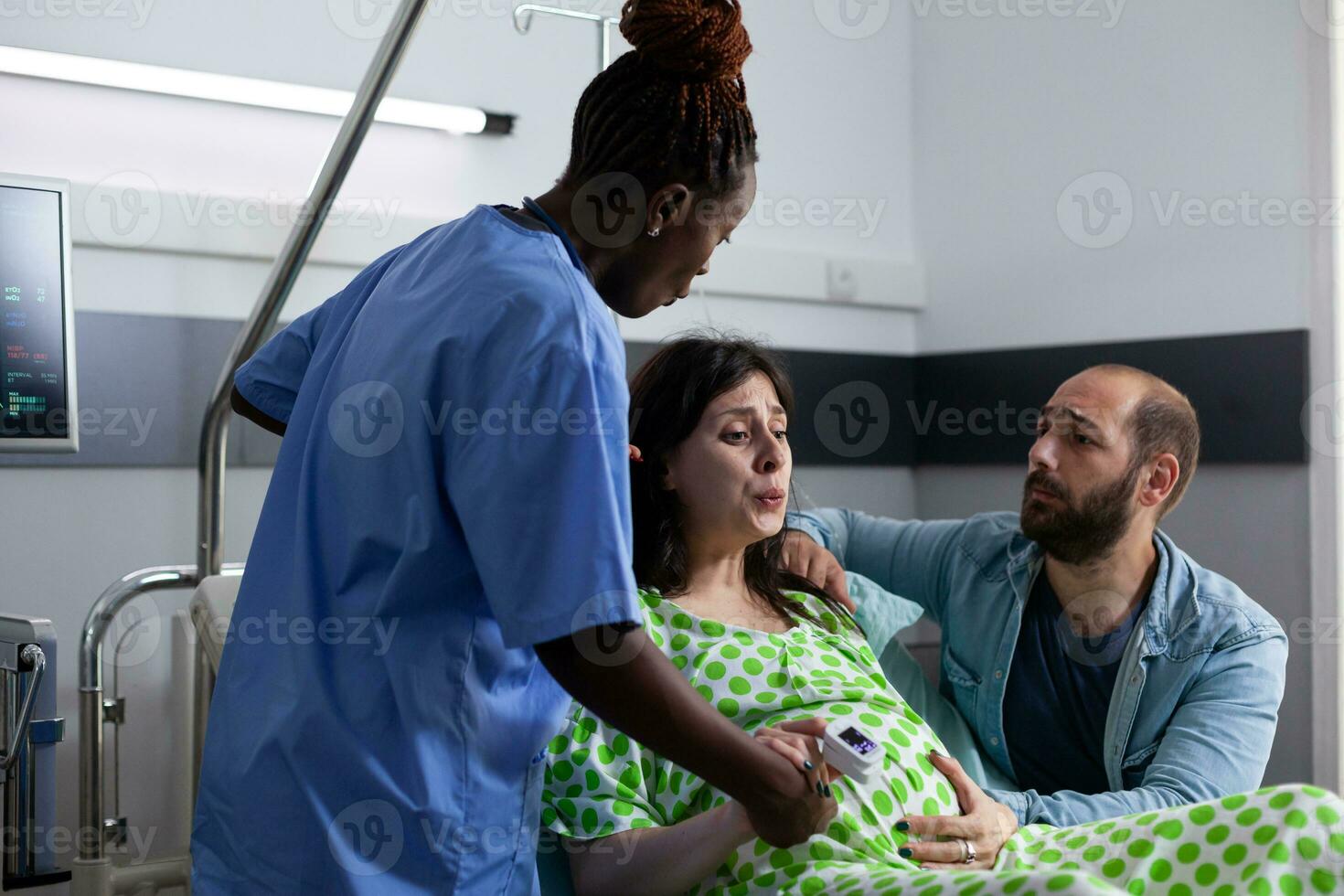 zwanger vrouw met pijnlijk arbeid aan het liegen in ziekenhuis afdeling bed, hebben weeën. Afrikaanse Amerikaans verpleegster en man ondersteunen toekomst moeder, Holding handen voor medisch bijstand foto