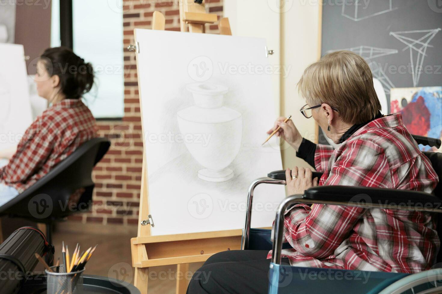 senior vrouw rolstoel gebruiker tekening met potlood Aan canvas, Bijwonen kunst klassen voor mensen met onbekwaamheid, verminderen spanning in creatief ruimte. creatief behandeling voor gehandicapt volwassenen foto