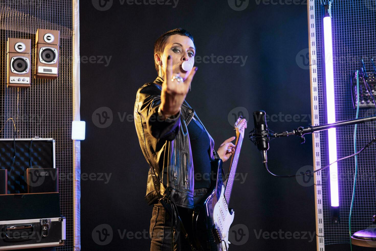 brunette gitarist met kort haar- aan het doen rockster gebaar Bij camera terwijl spelen elektrisch gitaar in studio, voorbereidingen treffen naar uitvoeren rots muziek- Bij metaal concert. performer blazen bubbel gom foto