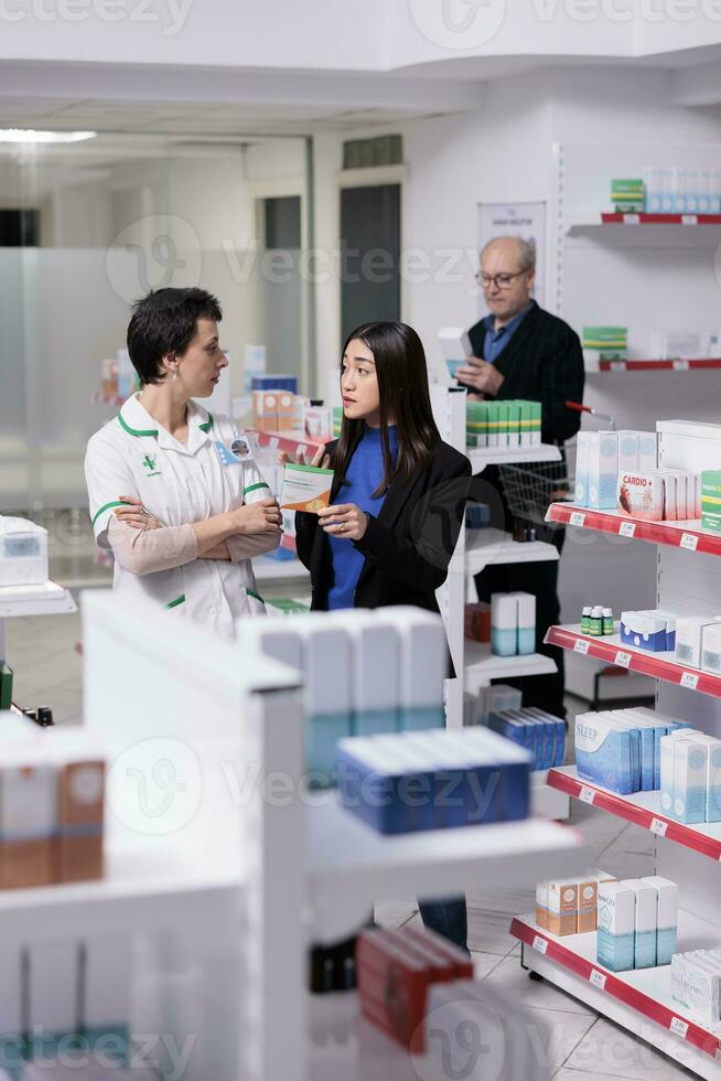 apotheek werknemer helpen vrouw kiezen vitamine c capsules in drogisterij. apotheker en jong Aziatisch klant staand in apotheek gangpad en bespreken ascorbinezuur zuur voordelen foto