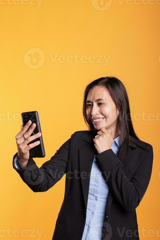 vrolijk Aziatisch vrouw pratend met afgelegen vriend gedurende video-oproep vergadering conferentie, hebben afgelegen discussie gedurende schieten breken in studio. volwassen in formeel pak staand over- geel achtergrond foto