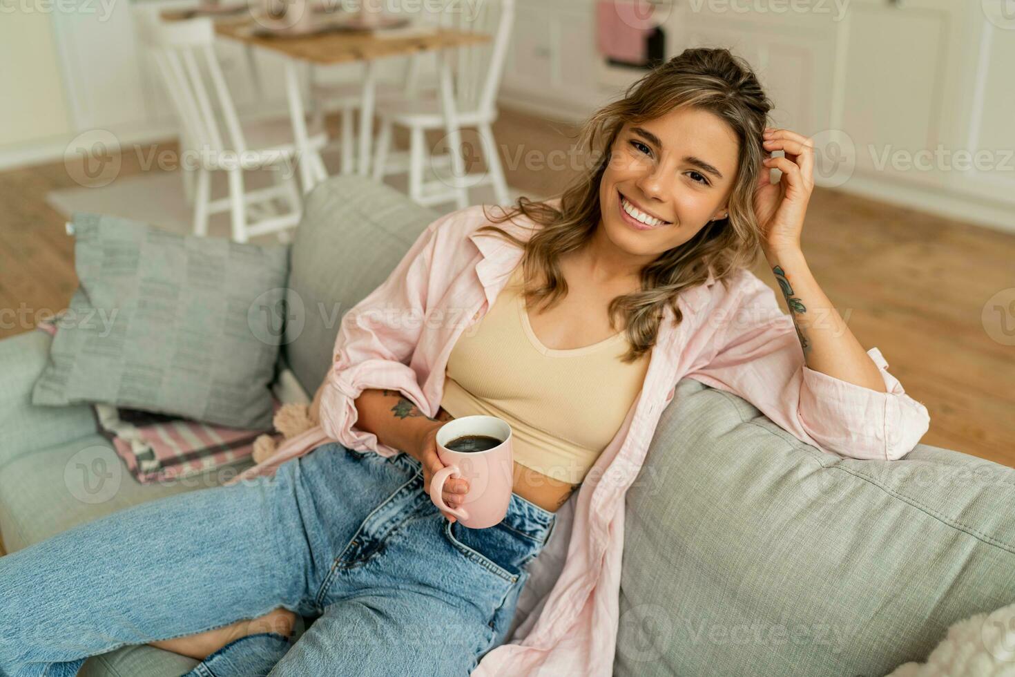 lief vrouw in gewoontjes kleren resting un sofa in knus leven kamer. Holding kop van cappuccino. foto