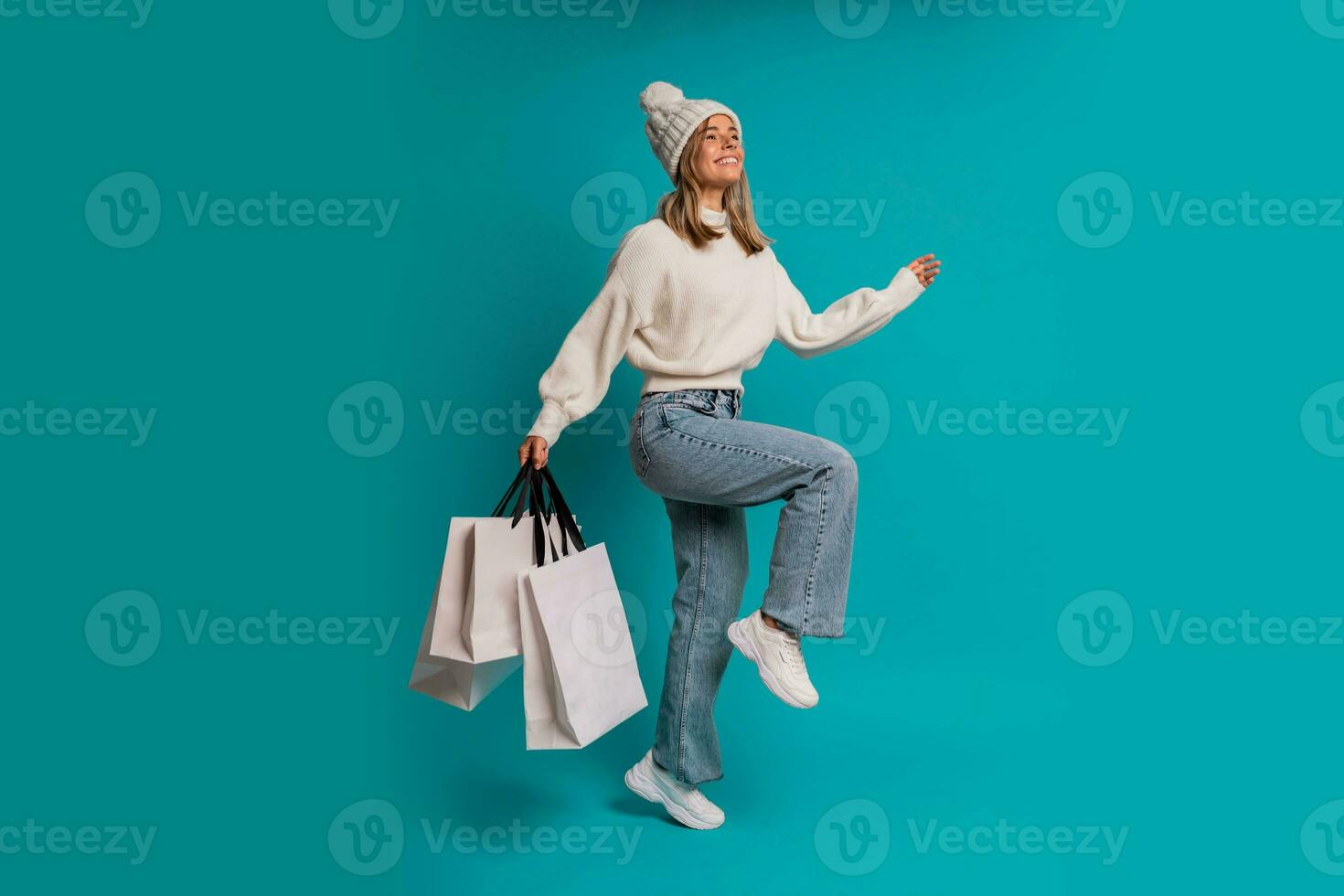 studio winter portret van glimlachen blond vrouw in wit hoed en whoo trui een Holding boodschappen doen Tassen , poseren in studio over- turkoois achtergrond. vol lengte. foto