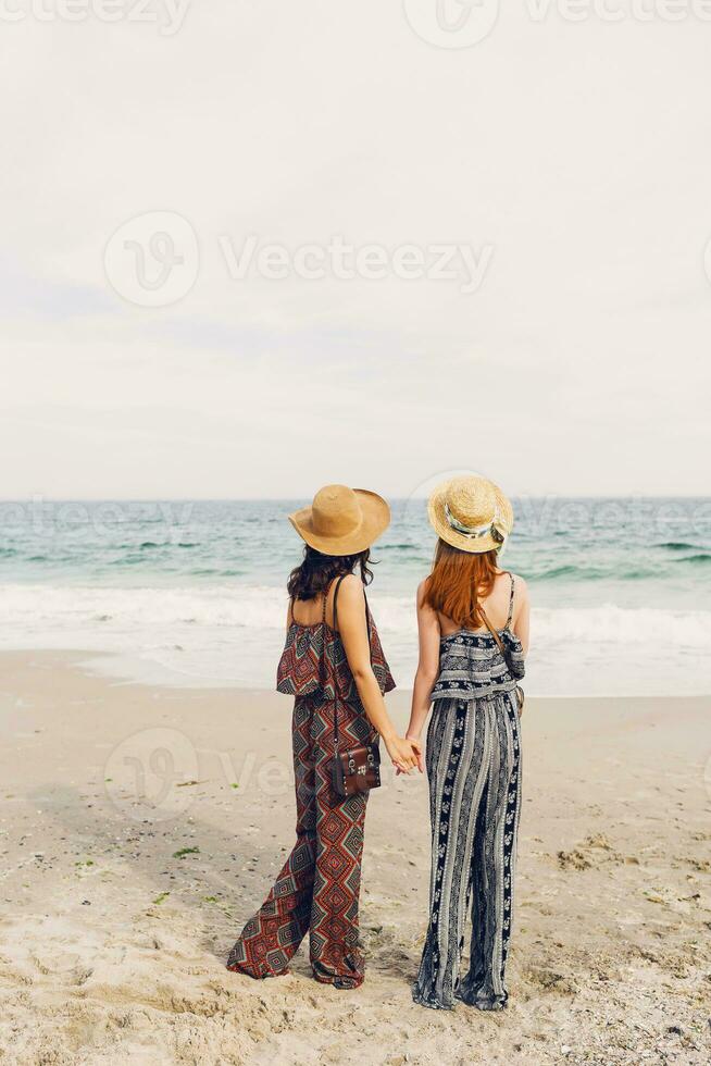 buitenshuis levensstijl beeld van twee meisjes het beste vrienden wandelen Aan de zee kust op zoek Bij camera lachend, praten. zussen wandelen langs een strand. elegant zomer strandkleding, rietje hoed , zonnig kleuren. foto