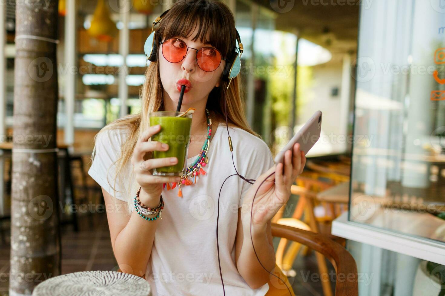 elegant vrouw in roze bril genieten van groen gezond smoothie , luisteren muziek- door oortelefoons, Holding mobiel telefoon. verrassing gezicht. modieus accessoires. tropisch humeur. foto