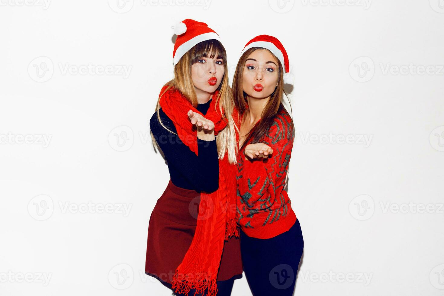 nieuw jaar feest. twee mooi meisjes in grappig maskerade de kerstman hoeden sturen kus . binnen- voorraad beeld van het beste vrienden poseren Aan wit achtergrond. isoleren. foto