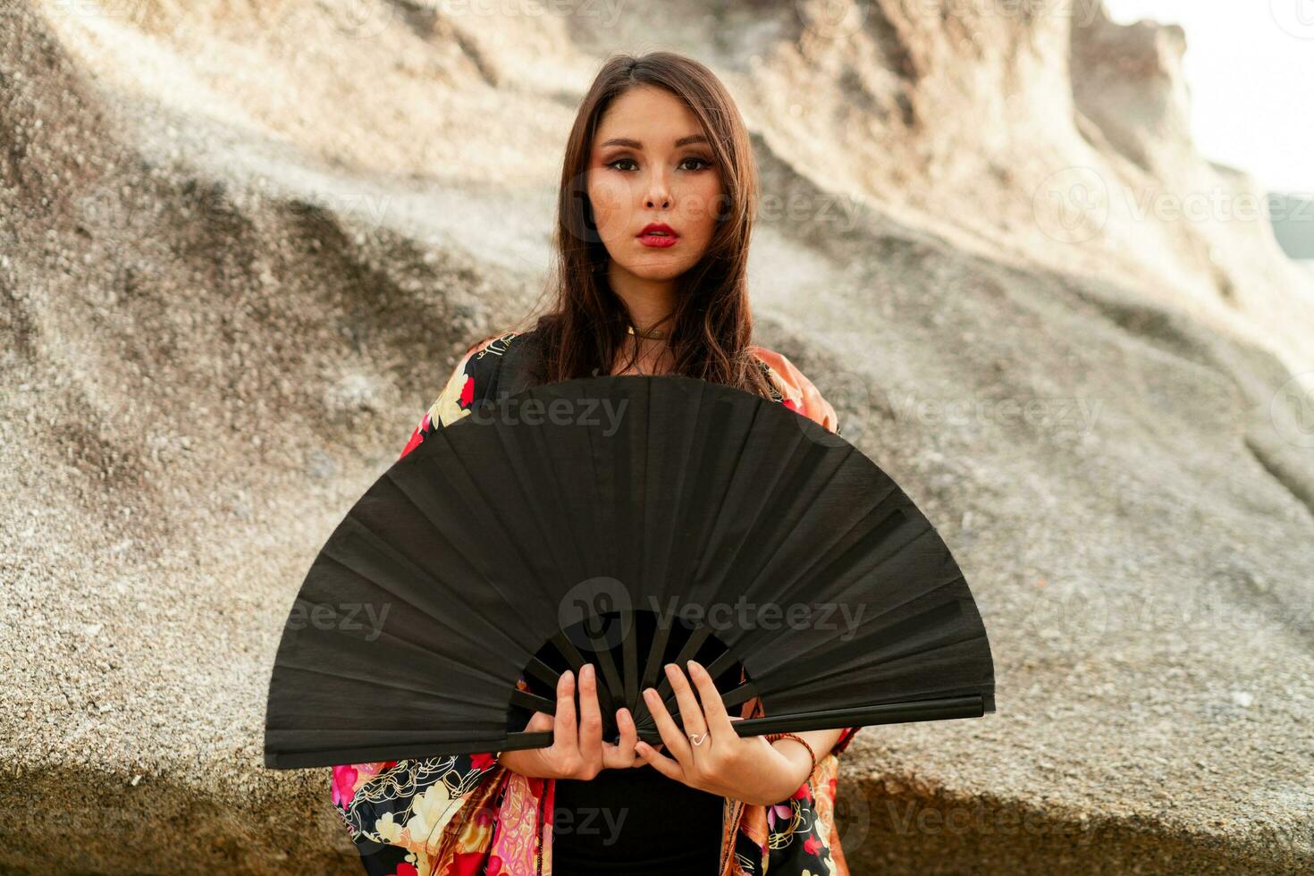 Aziatisch vrouw in kleurrijk zijde kimono blouse Holding ventilator en poseren Aan de strand met rotsen. foto