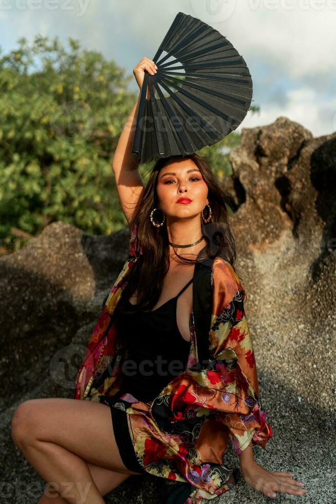 sensueel Aziatisch vrouw in zijde kimono Holding ventilator en poseren over- rotsen Aan de strand. foto