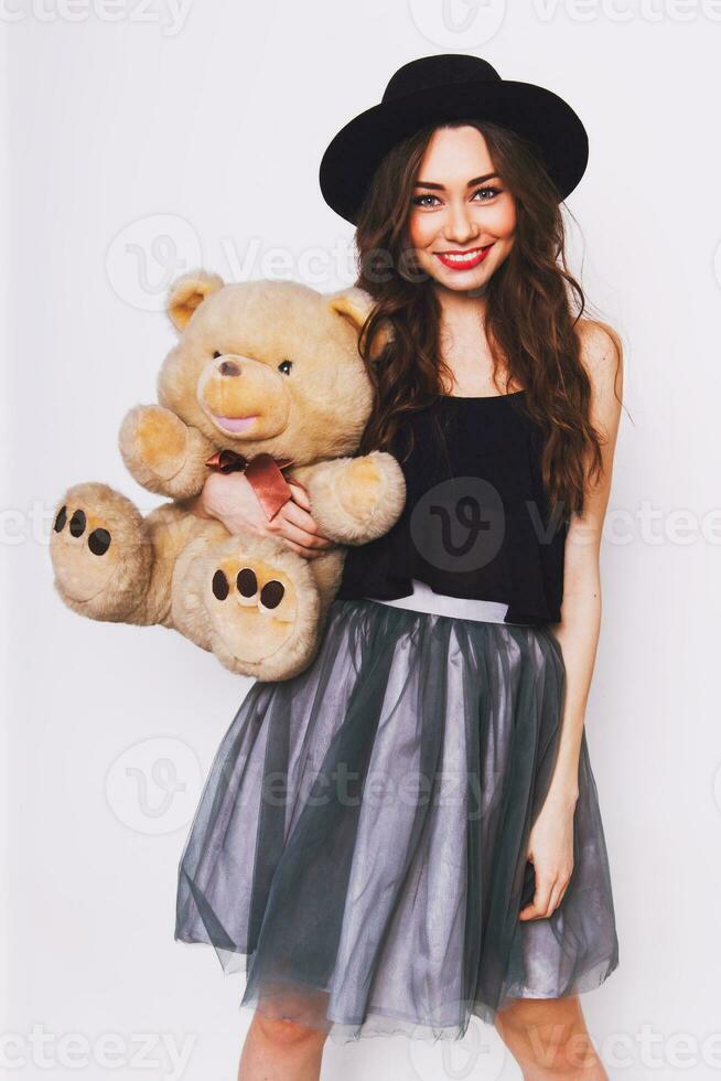 dichtbij omhoog portret van mooi schattig jong vrouw in elegant hipster kleding met groot ogen , rood lippen en perfect huid Holding haar pluizig teddy beer tegen wit stedelijk muur. foto