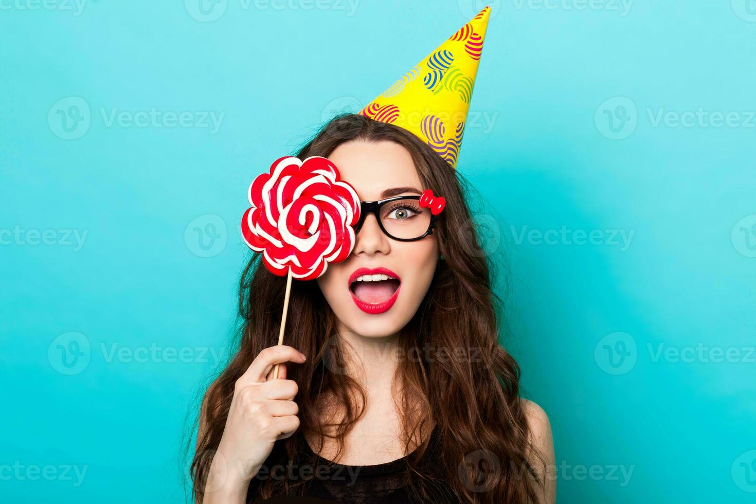 gek grappig mooi vrouw poseren tegen blauw muur vervelend modieus zomer kleding , koel schattig bril en partij hoed. Holding groot roze lolly . foto