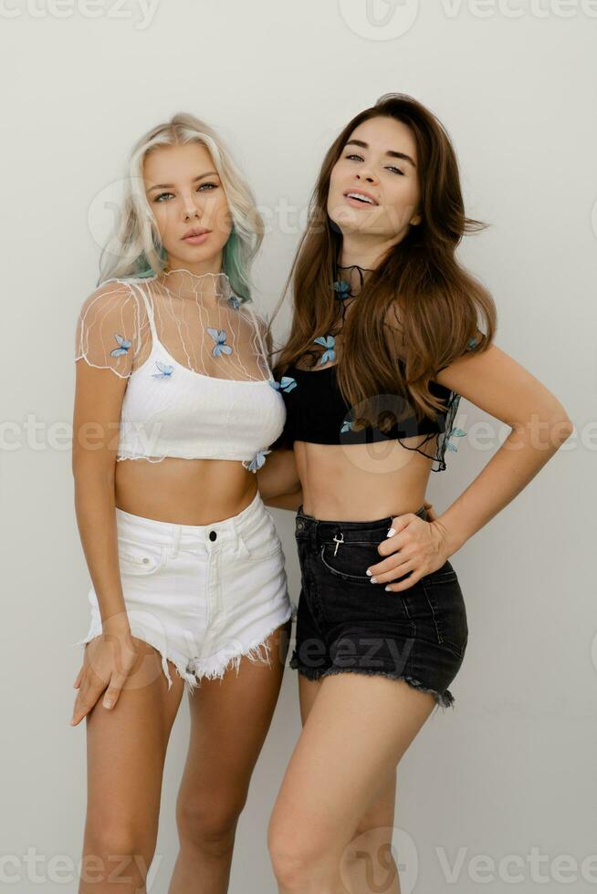 twee prachtig Dames met perfect haren in stijlvol zomer Bijsnijden tops met boterachtigen en jeans shorts poseren over- wit achtergrond. foto