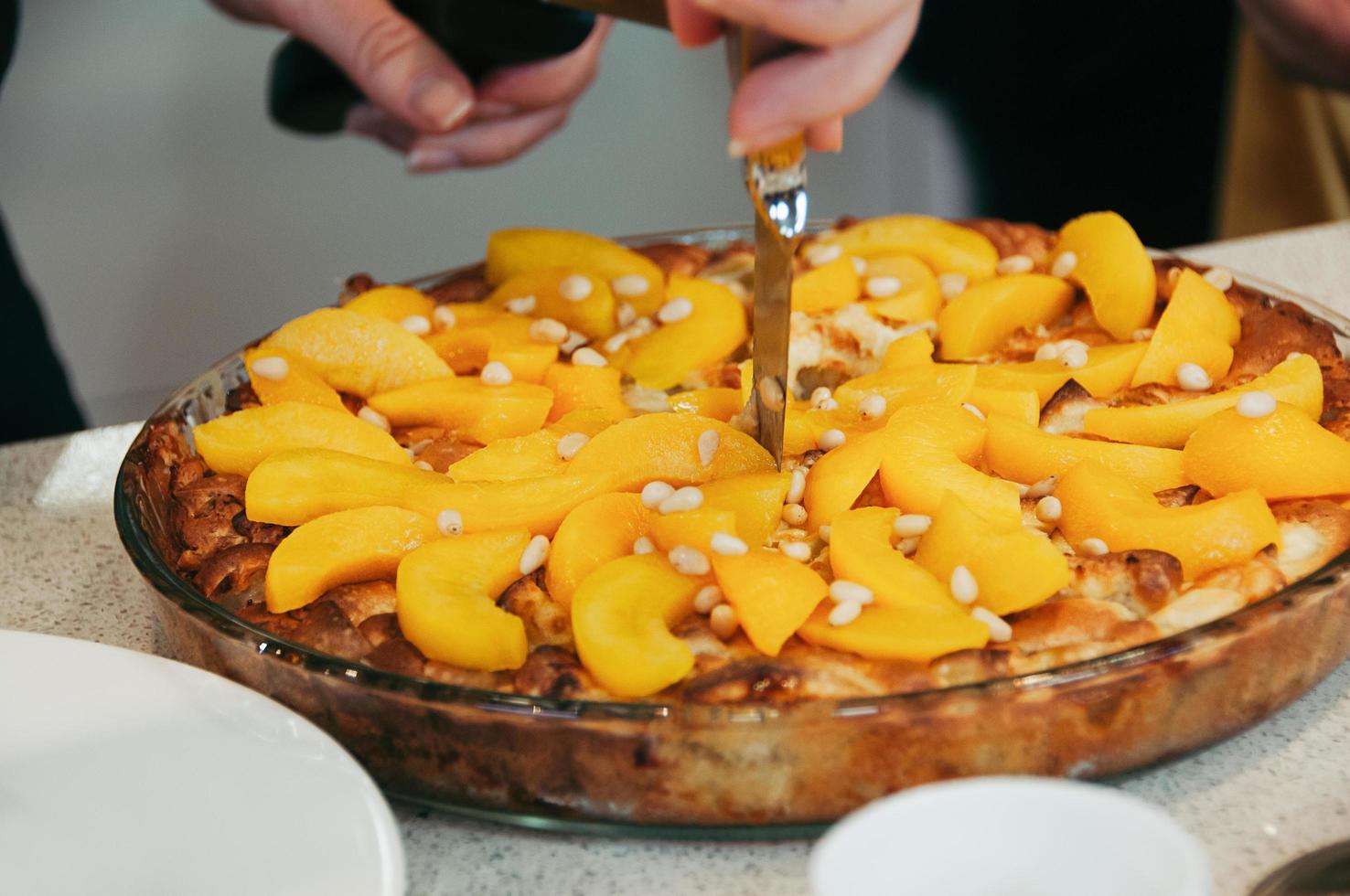 heerlijke veganistische perzikcake versierd met pijnboompitten, geheel, in ronde glazen pan, op tafel. selectieve aandacht. kleurrijke gezonde sappig. zijaanzicht, vrouwelijke hand met mes die de taart snijdt foto