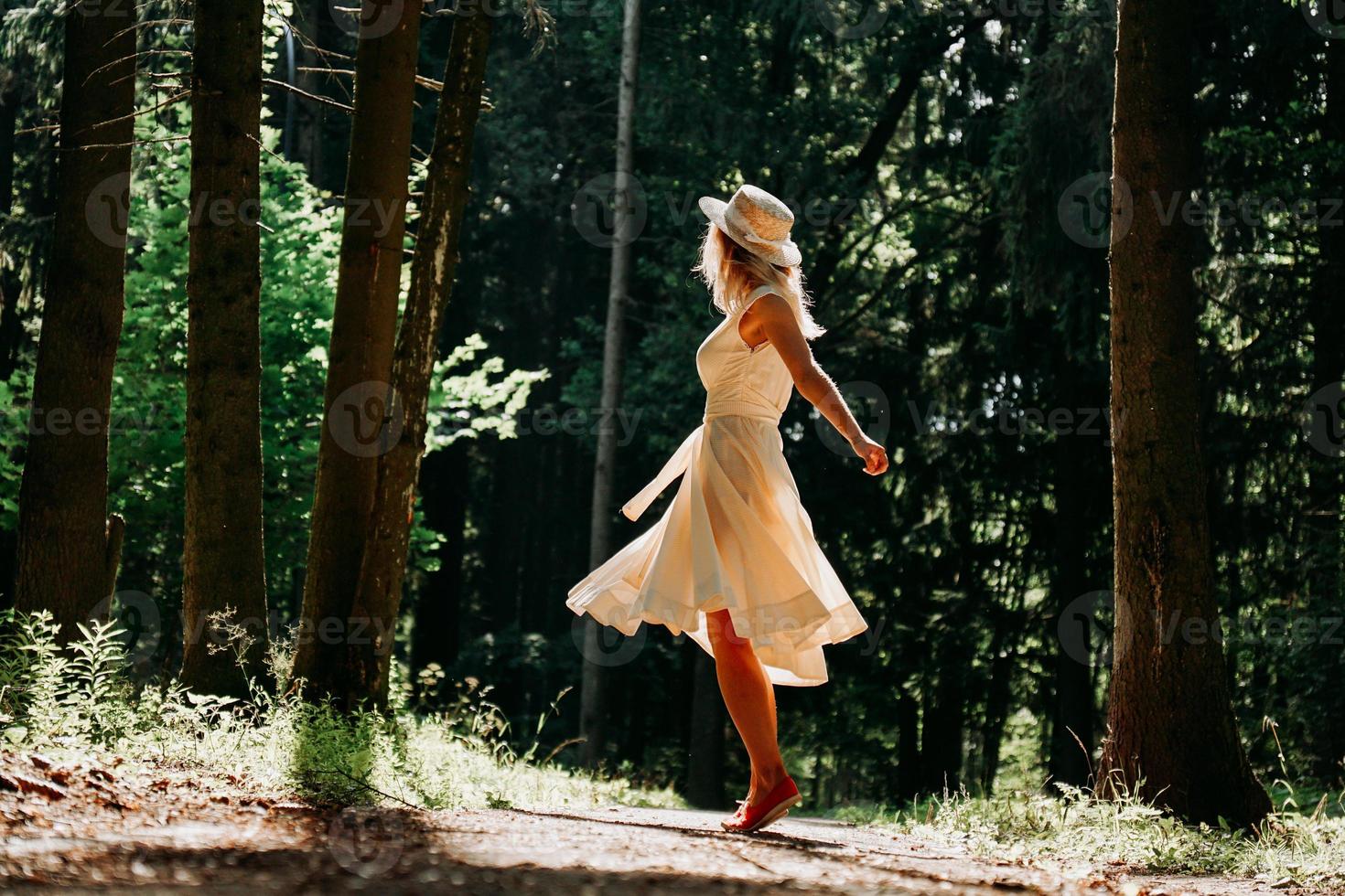 een jonge vrouw in een witte jurk en een strohoed loopt door het bos foto