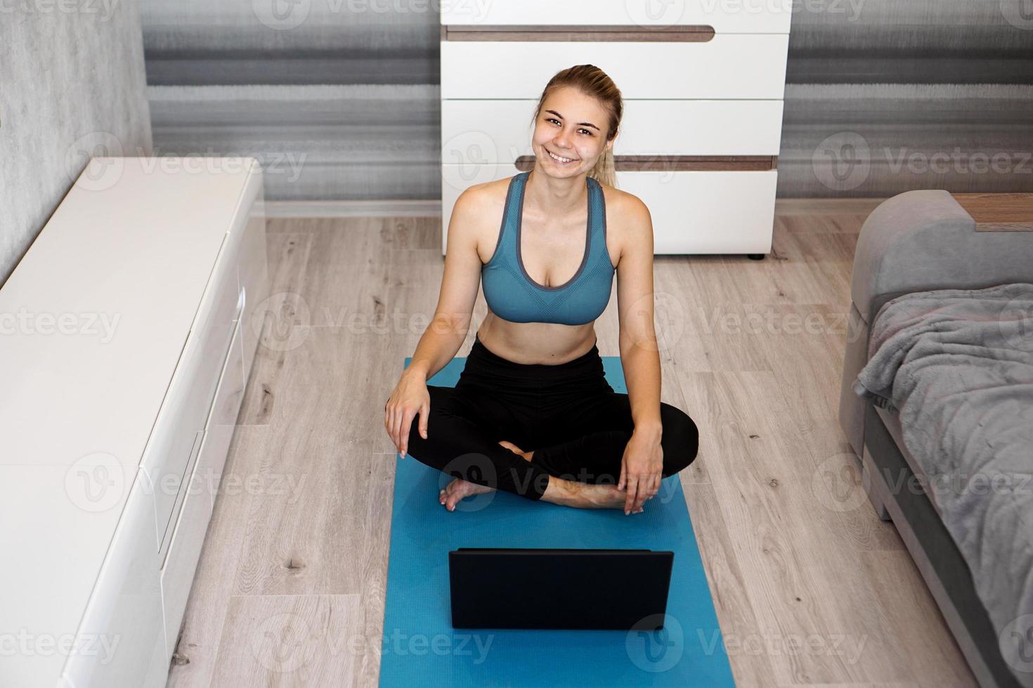 gelukkige jonge fitnessvrouw zit thuis op yogamat in de buurt van laptop foto