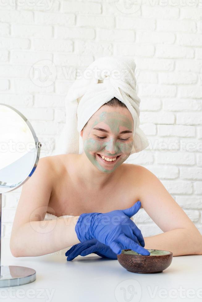 vrouw die handschoenen draagt die een gezichtsmasker van klei aanbrengt en in de spiegel kijkt foto