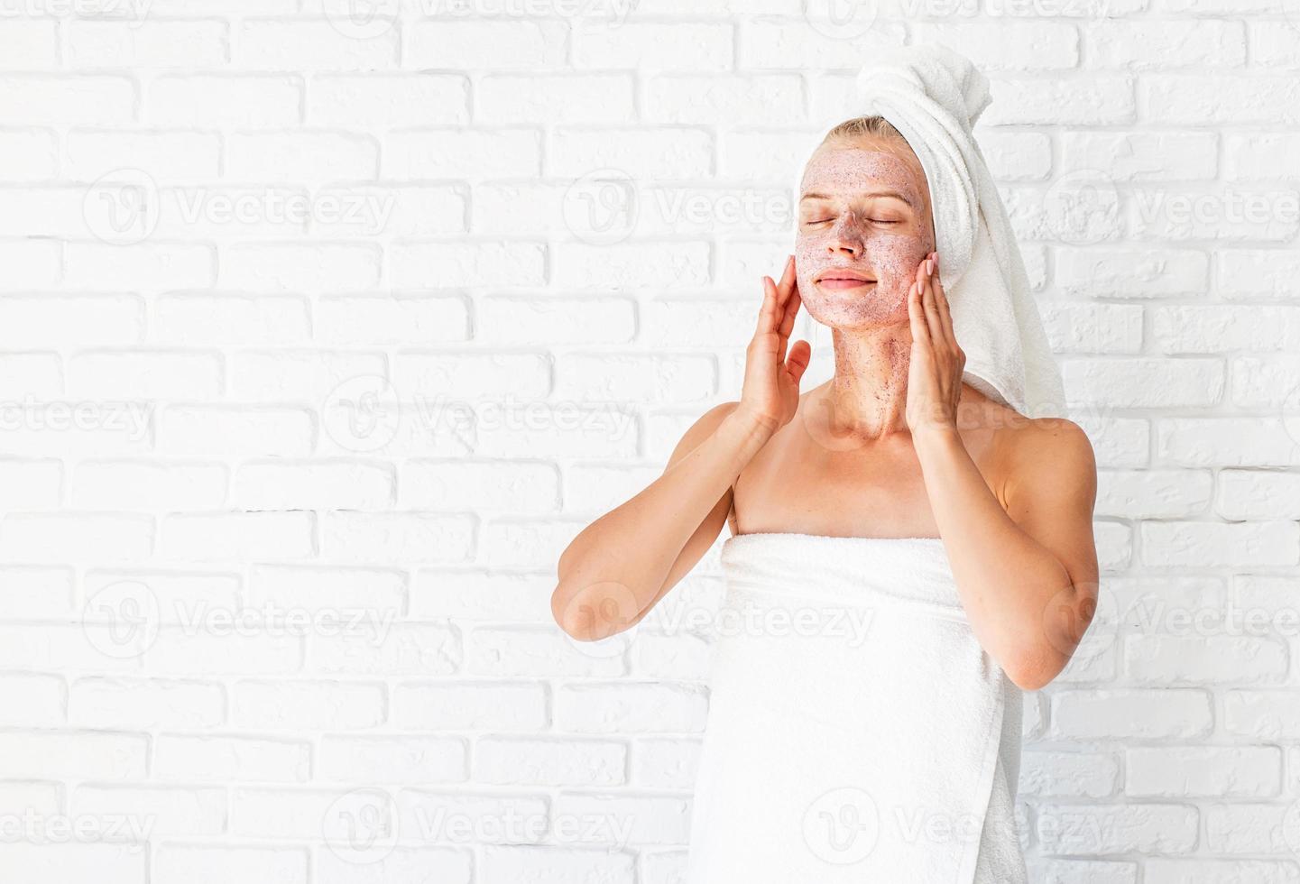 jonge vrouw in witte badhanddoeken die scrub op haar gezicht en nek aanbrengt foto