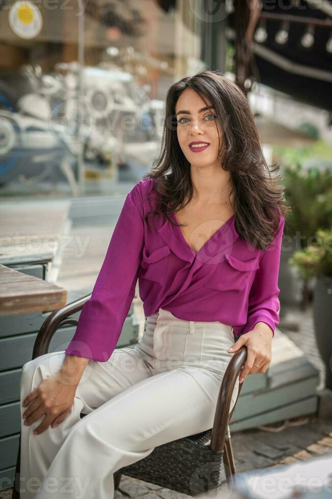 gelukkig brunette meisje met roze blouse zittend Bij de bar , drinken een glas van limonade terwijl glimlachen Bij de camera. jong sexy mooi vrouw Aan de stoel drinken sap vervelend een wit broek foto