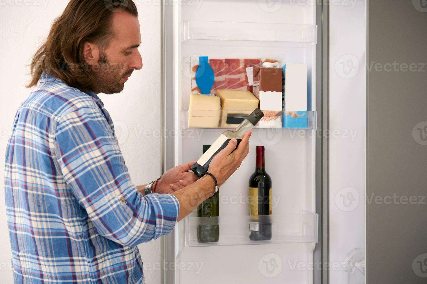 portret van een knap Mens Holding een fles van olijf- olie, staand in de buurt Open koelkast in de huis keuken foto